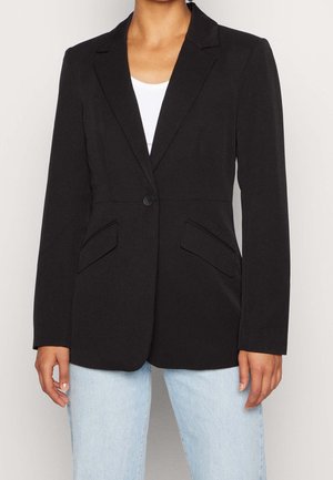Femme portant un blazer noir ajusté à un seul bouton sur un haut blanc et un jean bleu clair, debout devant un fond clair uni.