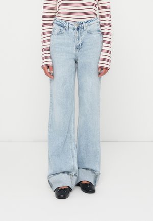 Persoon draagt lichtblauwe wijde jeans met opgerolde boorden, gestreept shirt met lange mouwen en zwarte ballerina's, staand op een witte vloer.