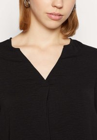 Blusa nera con un tessuto leggermente texturizzato. Presenta un scollo a V ed è progettata con una vestibilità comoda, che mette in risalto uno stile minimalista.