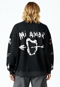 Sweatshirt noir surdimensionné avec un texte graphique blanc "Mi Amor", flèches, designs de crâne et de cœur au dos et sur les manches ; poignets et ourlet côtelés.