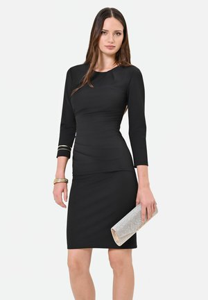 Donna con un abito nero aderente a maniche tre quarti, che tiene una pochette argentata e indossa braccialetti dorati a un polso.
