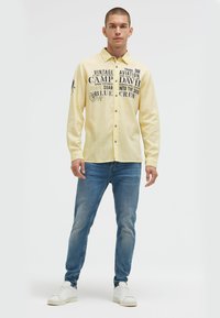 Helles gelbes Langarmhemd mit bedrucktem Text und Aufnähern, kombiniert mit blauen Jeans und weißen Sneakers. Lässige Passform und mit Knöpfen zu schließen.