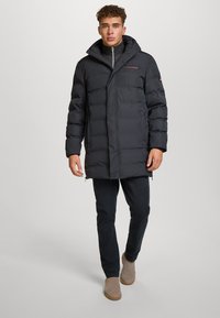 Polstret svart parkajakke med høy krage, glidelåslommer og logo på brystet. Båret med mørke bukser og beige støvler.