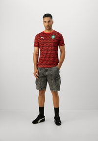 Rød mønstret kortærmet t-shirt med rund hals, der har et grønt logo. Sammen med grå cargo shorts og sorte sneakers.