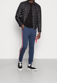 Schwarze Pufferjacke, schwarzes T-Shirt, marineblaue Jogginghose mit roten Seitenstreifen und schwarze Sneakers mit weißen Sohlen. Einfaches, lässiges Outfit.
