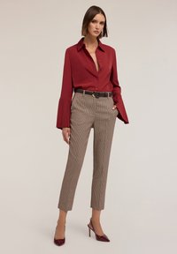 Camicia rossa con bottoni e maniche lunghe plissè, abbinata a pantaloni marrone bicolore a zigzag e scarpe con tacco bordeaux. Cintura nera con fibbia dorata.