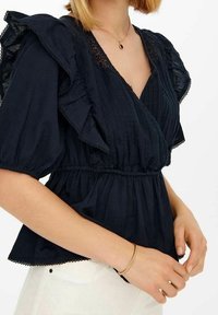 Blusa blu navy con maniche a volant, texture plissettata e rifinitura in pizzo. La vita è elasticizzata per una silhouette aderente. Accentuata da un delicato motivo.