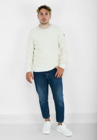 Maglione bianco a coste con scollo rotondo, abbinato a jeans in denim blu e sneakers bianche. Fit semplice e moderno.