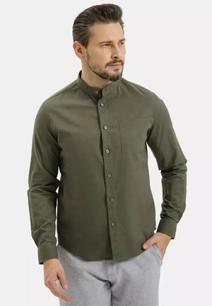 Mężczyzna w oliwkowej koszuli z kołnierzem typu mandarynka, zapinanej na guziki z długimi rękawami i szarych spodniach, patrzy w prawo z jedną ręką w kieszeni.