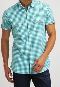 Camisa de manga corta a cuadros turquesa y blancos con cierre de botones, dos bolsillos en el pecho y puños remangados, confeccionada con tela ligera.