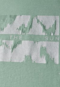 Mintgrøn T-shirt med et tekstureret, slidt hvidt grafisk design, hvor ordene "THE SUN" er integreret i mønsteret.