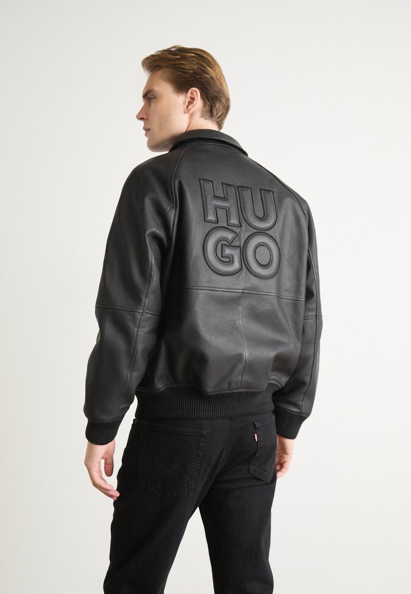 Melnā ādas bumbas jaka ar ribotām piedurknēm un apakšmalu. Aizmugurē ir liels izspiedums "HUGO" logo. Modelis ir nedaudz pagriezies, attēlojot dizainu.