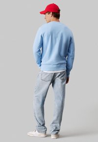 Ανοιχτό μπλε φούτερ με ριχτές μανσέτες και στρίφωμα, συνδυασμένο με ξεθωριασμένα μπλε τζιν και λευκά αθλητικά παπούτσια. Κόκκινο καπέλο φορεμένο ανάποδα.