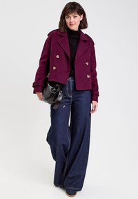 Femme portant un manteau court violet à double boutonnage, un col roulé noir, un jean large foncé et tenant un sac en cuir noir.