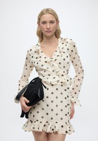 Ξανθιά γυναίκα με διάφανο μπεζ φόρεμα με μαύρες πουά και βολάν κρατά μια μαύρη τσάντα χειρός σε λευκό, απλό φόντο.