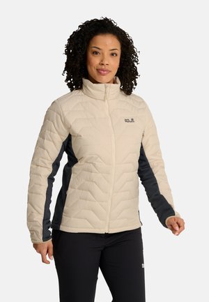 Donna con capelli ricci che indossa una giacca beige e nera con zip e pantaloni neri, in piedi davanti a uno sfondo chiaro e uniforme.