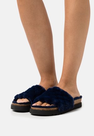 Mules en fourrure bleu marine avec un design texturé et matelassé. Semelle intérieure en liège et semelle extérieure en caoutchouc offrant confort et stabilité. Style ouvert.