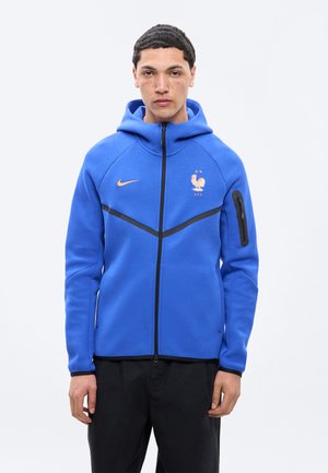 Jonge man die een blauwe hoodie met rits draagt, met het Nike-logo en het Franse voetbalembleem, staand tegen een effen lichte achtergrond.
