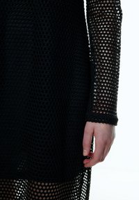 Schwarzes Mesh-Kleid mit langen Ärmeln und lockerer Passform. Verfügt über ein sechseckiges Muster für Atmungsaktivität. Der Saum endet bis zur Mitte des Oberschenkels.
