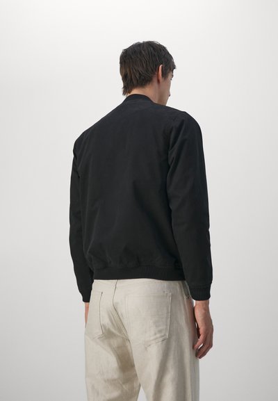 Chaqueta bomber negra con cuello y puños de canalé, con una textura suave. Llevada con pantalones de color claro, vista desde la parte trasera.