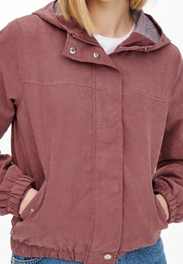Personne portant une veste à capuche mauve avec des boutons-pression et des poignets élastiques, les mains dans les poches latérales, sur un jean bleu et une chemise blanche.