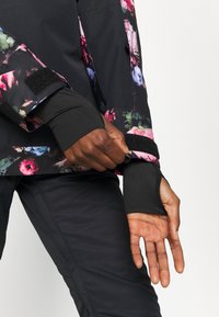 Giacca nera con motivo floreale e maniche aderenti, dotata di polsini in velcro. Indossata sopra pantaloni neri, che mostrano una texture di tessuto liscia.