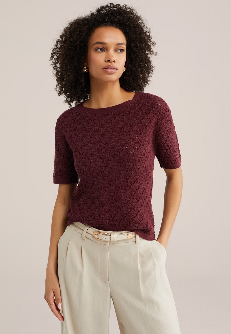 Lokkis juustega naine kannab tekstuuriga lühikeste varrukatega burgundia punast kampsunit ja kõrge vöökohaga beeže pükse heleda vööga, seisab käsi taskus.