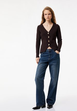 Vrouw die een zwarte, knoopsgat V-hals cardigan en losse blauwe spijkerbroek draagt, staand met één hand in de zak tegen een effen achtergrond.