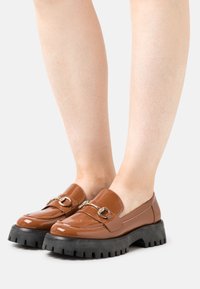 Mocassins en cuir verni marron avec une semelle en caoutchouc noir épais, dotés de quincaillerie décorative en ton doré et d'une finition lisse.