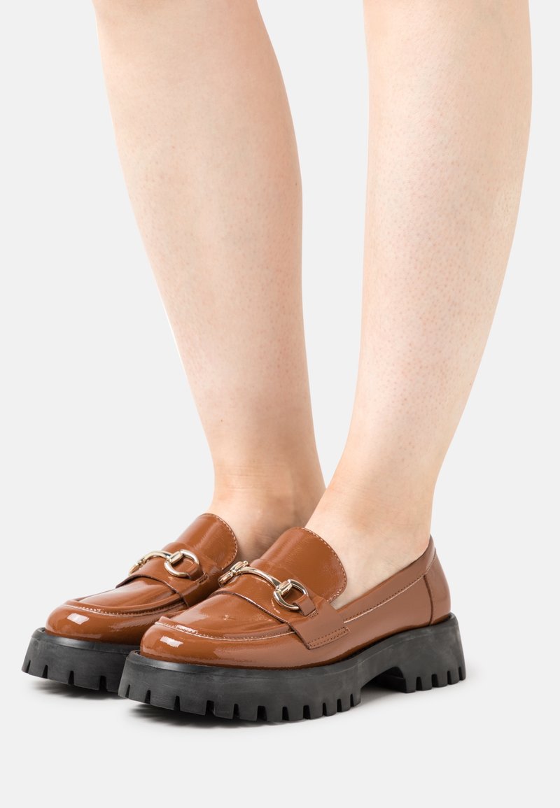 Mocassins en cuir verni marron avec une semelle en caoutchouc noir épais, dotés de quincaillerie décorative en ton doré et d'une finition lisse.