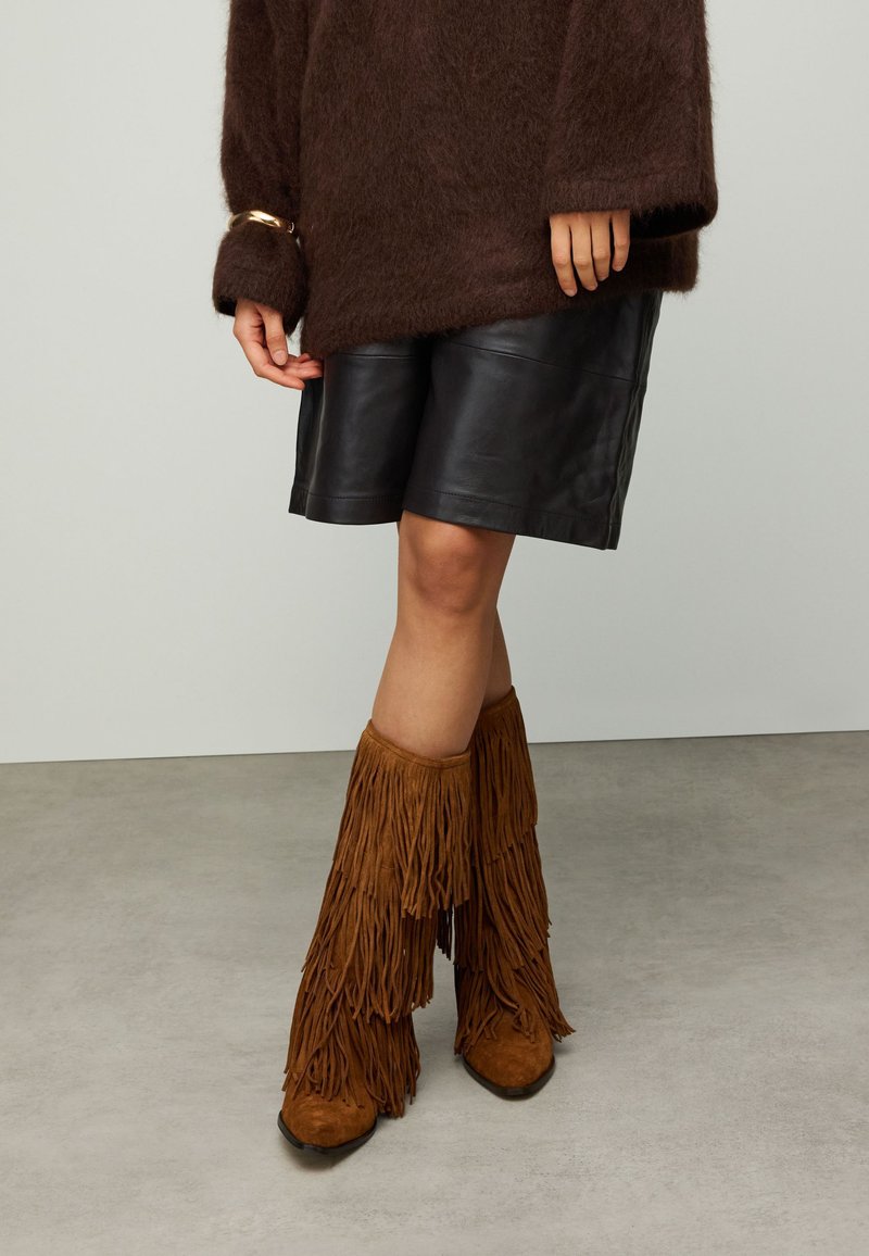 Brauner flauschiger Pullover, schwarze Leder-Shorts und braune kniehohe Stiefel mit Fransen. Der Pullover hat weite Ärmel; die Stiefel sind mit einer mehrlagigen Fransenoptik gestaltet.