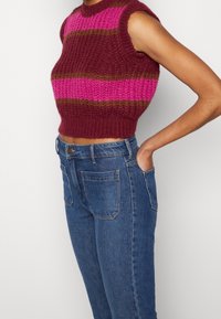 Haut court en maille sans manches avec des rayures horizontales rose, bordeaux et marron, porté avec un jean bleu taille haute, main dans la poche.