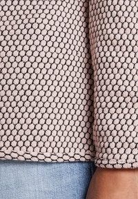 Stickad tröja i mjuk rosa med svart vävd mönster. Har en texturerad yta och en detalj vid nederkanten, visad mot ljus denimtyg.