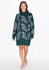 Oversized türkisfarbener Strickkleid mit silbernen Blättermustern, hohem Kragen und langen Ärmeln; getragen mit hellen Ballerinas.