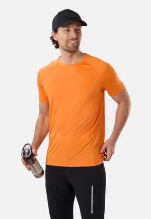 Mann in orangefarbenem Sportshirt, schwarzer Mütze und schwarzer Hose, hält eine transparente Wasserflasche und blickt lächelnd seitlich.
