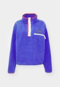 Pullover in pile di colore blu brillante con tasca con zip, colletto alto con accenti rossi e pattina con bottoni bianchi; superficie testurizzata.