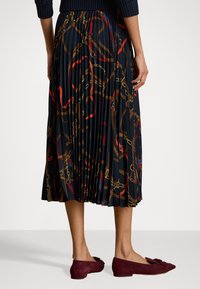 Fustă plisată din țesătură navy cu un imprimeu cu lanțuri de aur și panglici. Asortată cu balerini burgundy, accentuați de un detaliu cu fundiță pe vârf.