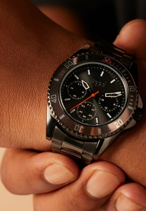 Schwarze Metallarmbanduhr mit drei Unterzifferblättern und orangefarbenem Sekundenzeiger an einem Handgelenk mit mittlerem braunem Hautton.