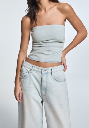 Kvinde iført lysegrå stropløs top og løstsiddende lyseblå jeans med den ene hånd på hoften mod ensfarvet baggrund.