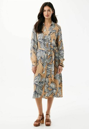 Kobieta ubrana w beżową sukienkę midi z długim rękawem, ozdobioną dużymi białymi i czarnymi kwiatowymi wzorami, z wiązanym w talii paskiem oraz brązowymi sandałami na platformie.