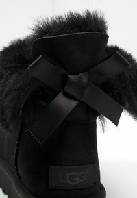 Bottine chevilles en daim noir avec un col en fourrure douce, nouée avec un ruban en satin noir. Le logo UGG est visible sur le talon.