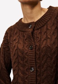 Brauner kabelgestrickter Pullover mit dunklen Knöpfen, getragen von einer Person, Nahaufnahme des Oberkorpus und des Ausschnitts.
