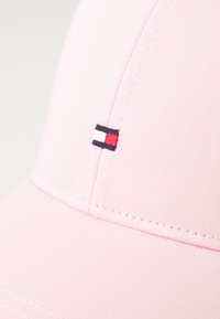 Rosa tygkeps med en sydd logotyp med blå, vita och röda ränder. Designen inkluderar en rundad skärm och textilmaterial med struktur.
