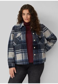 Frau mit langen lockigen Haaren, die eine blau-grau karierte Jacke über einem bordeauxroten Rollkragenpullover und schwarzen Hosen trägt, steht vor einem grauen Hintergrund.