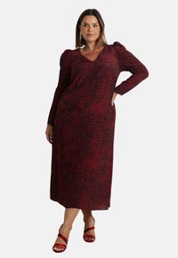 Maxi šaty s leopardím vzorem v červené a černé barvě, dlouhými rukávy, výstřihem do V, detaily na ramenou a kapsami. Skombinováno s červenými sandály na tenkých páscích.