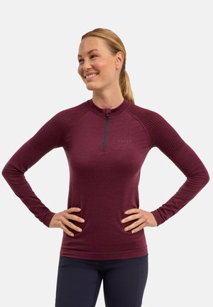 Frau mit blondem Pferdeschwanz, die ein langärmeliges bordeauxrotes Sporttop mit strukturierten Schultern und einem Frontreißverschluss trägt, lächelt mit den Händen in der Hüfte.