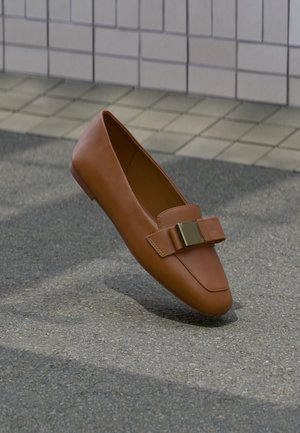Chaussure plate en cuir marron avec une boucle dorée décorative à l'avant, flottant au-dessus d'un sol en béton et carrelé.