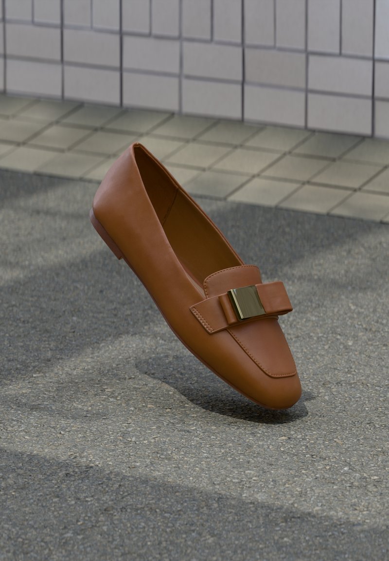 Chaussure plate en cuir marron avec une boucle dorée décorative à l'avant, flottant au-dessus d'un sol en béton et carrelé.