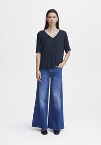 Černý top s výstřihem do V a krátkými rukávy; kombinovaný s volnými modrými džíny. Modelka stojí s jednou rukou v kapse, minimalistické šperky jako doplněk.