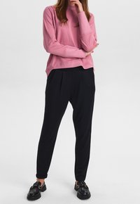 Rosa Sweatshirt mit lockerer Passform und langen Ärmeln, kombiniert mit schwarzen, gefältelten Hosen. Die Schuhe haben ein glänzendes schwarzes Finish und eine Schnallen-Details.